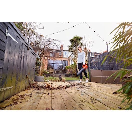 BLACKDECKER BCBL200L leaf blower held by person clearing leaves on wooden deck in garden.