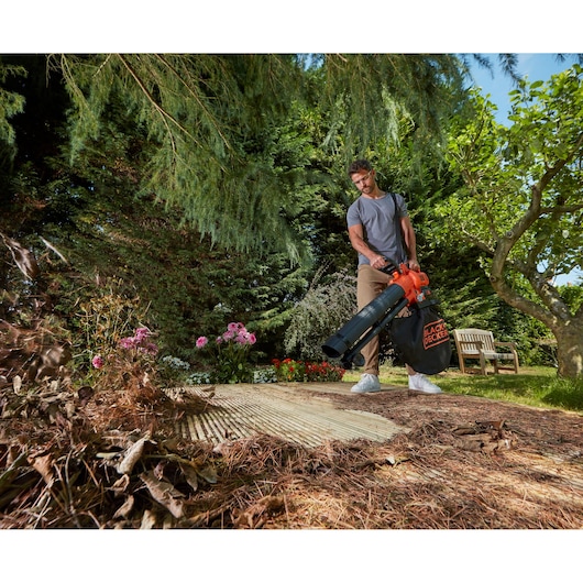 Person using BLACK+DECKER BCBLV36B leaf blower vacuum in a garden clearing dried leaves.