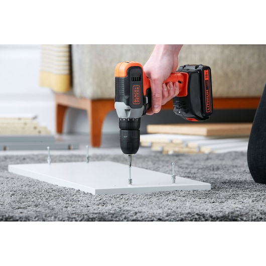 BLACK+DECKER BCD001C1 cordless drill in use driving screws into a white wooden board on a carpeted floor.