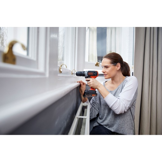 Person using BLACK+DECKER BCD001C1 cordless drill on a window frame indoors.
