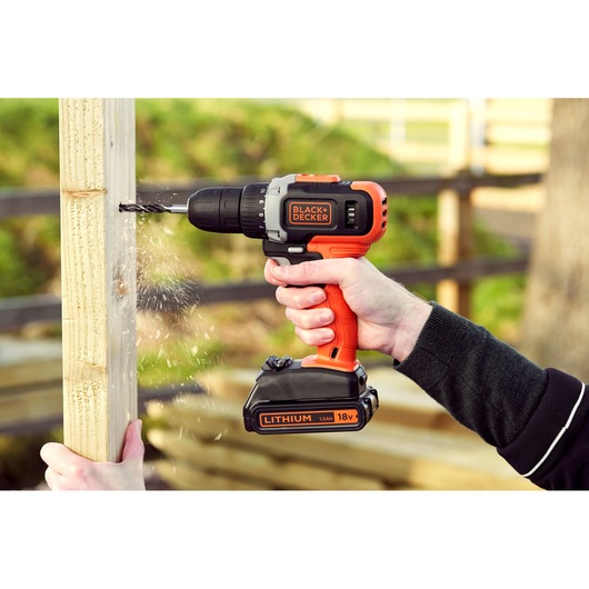 BLACK+DECKER BCD002C1 cordless drill in use drilling wood, orange and black with visible lithium 18V battery.