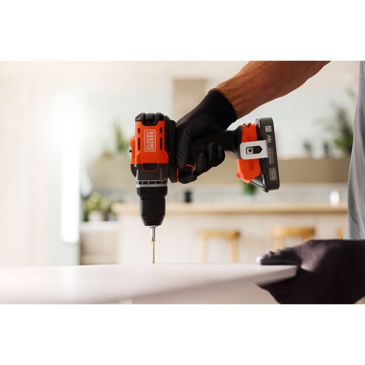 A BLACK+DECKER cordless drill is used to drive a screw into a white surface in a modern indoor setting.
