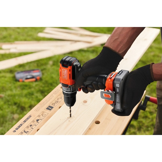 Cordless BLACK+DECKER drill in use, drilling into wood outdoors. Person wearing gloves holds the tool.
