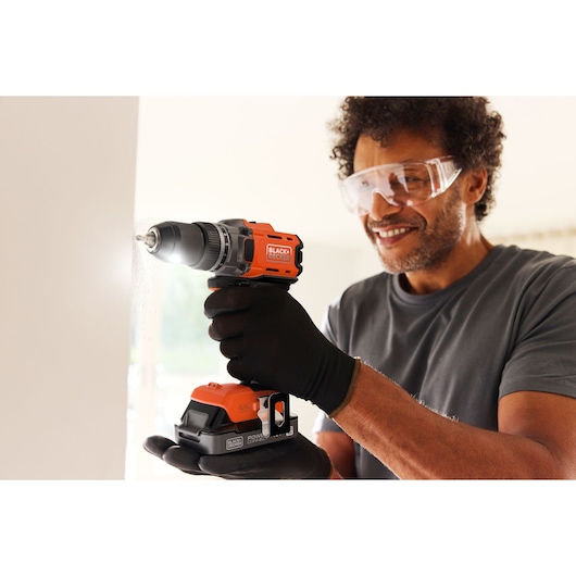 Person using a BLACK+DECKER cordless drill to make a hole in a wall, wearing safety glasses and gloves.