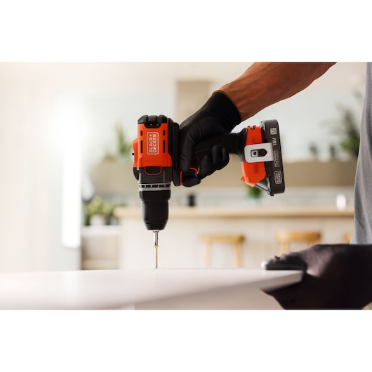 BLACK+DECKER cordless drill in use, drilling a screw into a white surface in a modern indoor setting.