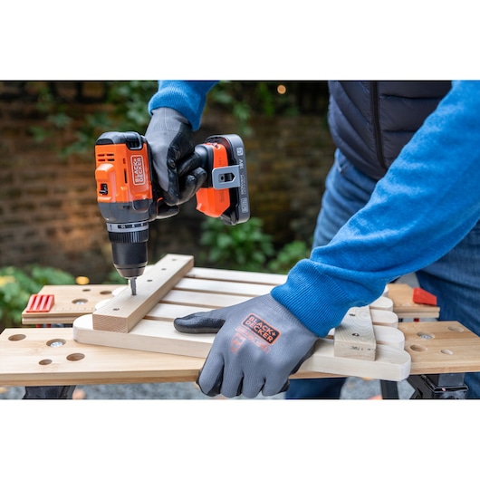 Person using BLACK+DECKER cordless drill to assemble wooden pieces on a workbench outdoors.
