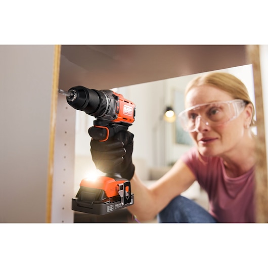 Person using a BLACK+DECKER cordless drill to assemble furniture, wearing safety glasses and gloves.