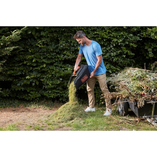 Photo of a person emptying a BLACK+DECKER BCMW3336L1 lawn mower grass bag outdoors onto a pile of clippings.