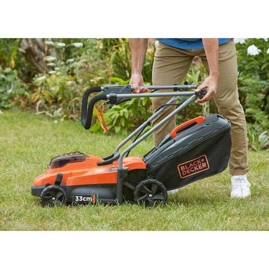 Photo of BLACK+DECKER BCMW3336N lawn mower with black and orange design, foldable handle, and grass collection box in use on grass.
