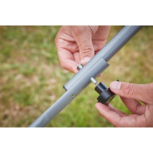 Close-up of hands assembling BLACK+DECKER BCMW3336N metal tube and knob outdoors, grass background.