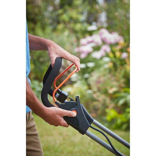 Photo of a hand gripping the BLACK+DECKER BCMW3336N lawn mower handle with orange safety bar, outdoor garden background.