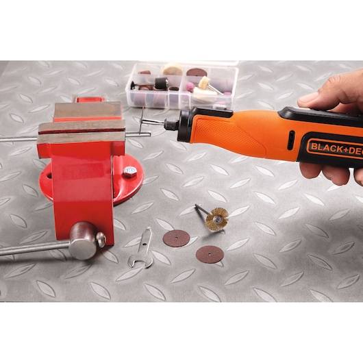 BLACK+DECKER BCRT8IK rotary tool in use, held near a red vise with accessories and tools on a metal work surface.