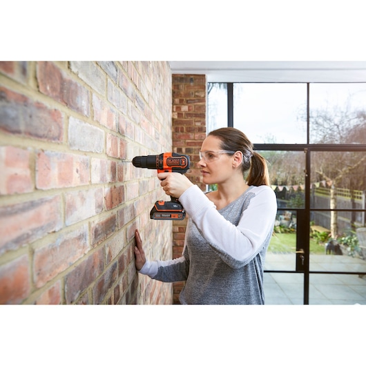 Person using BLACKDECKER BDCHD18S1KA cordless drill to drill into indoor brick wall, garden visible through window.