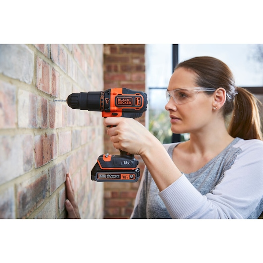 Photo of BLACK+DECKER BDCHD18 cordless drill used on brick wall, black and orange, held by person.