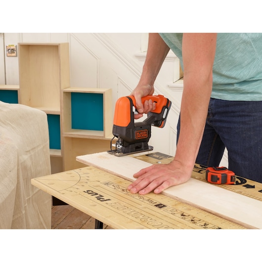BLACKDECKER BDCK123S2S cordless jigsaw cutting wood on workbench, orange and black tool in use.