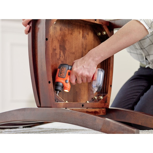 A person using an orange and black BLACK+DECKER cordless screwdriver to assemble or repair a wooden piece of furniture.