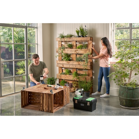 BLACKDECKER BDST60120 black toolbox with green lid beside wooden planters and plants in a bright indoor setting.