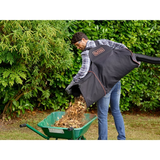 BLACKDECKER BEBLV300 garden vacuum bag in use, emptying mulch into a green wheelbarrow outdoors.