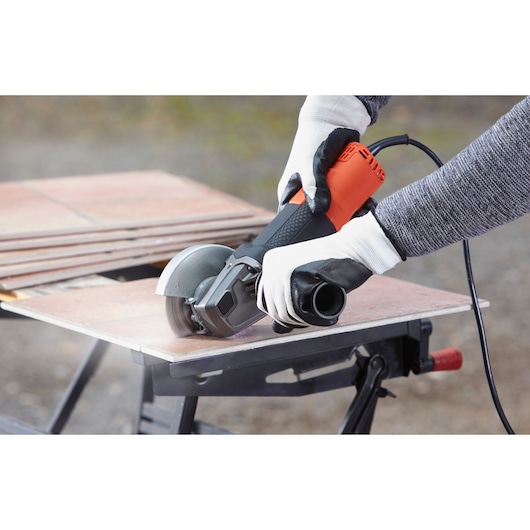 BLACKDECKER BEG210K angle grinder cutting tile on workbench, orange and black body held with gloves.