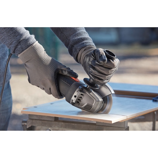 BLACKDECKER BEG220K angle grinder in use cutting tile, close-up of hands wearing gloves outdoors.