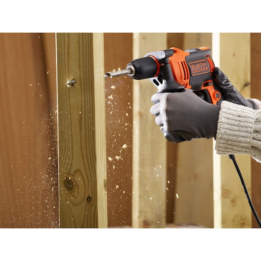 BLACK+DECKER BEH710 corded drill in use drilling through wood, close-up view, showing sawdust and gloved hands.