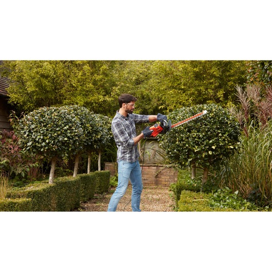 Person trimming bushes with BLACKDECKER BEHTS301 hedge trimmer in a garden.