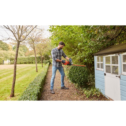 Person trimming hedge outdoors with BLACKDECKER BEHTS301 hedge trimmer next to a blue garden shed.