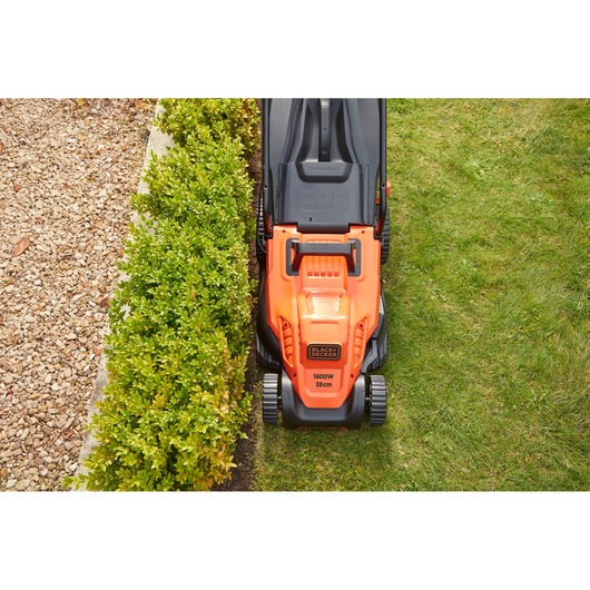 Photo of BLACKDECKER BEMW471BH orange lawn mower cutting grass next to green hedge and gravel path.