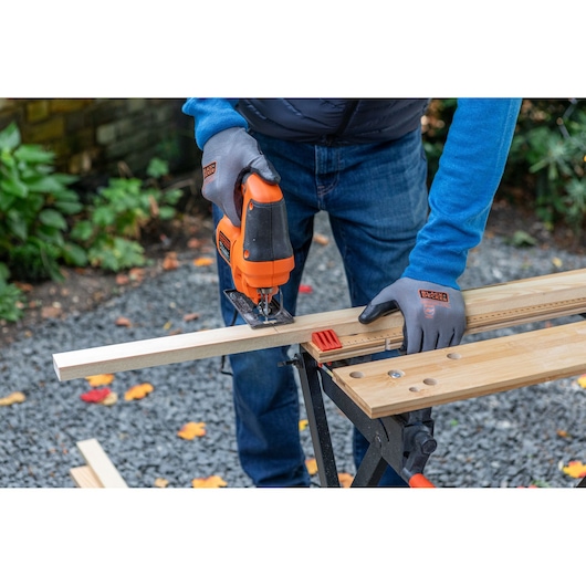 BLACKDECKER BES602 jigsaw in use, cutting wood on outdoor workbench, user wearing branded gloves.