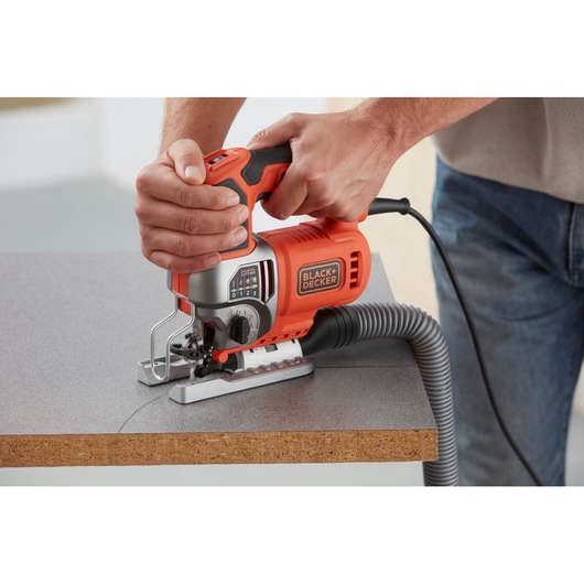 Photo of BLACK+DECKER BES610 jigsaw cutting a wood board, orange and black power tool with dust extraction hose
