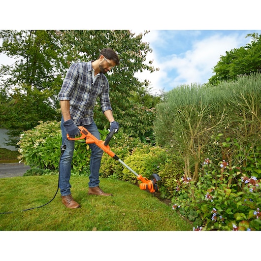 Photo of person using BLACKDECKER BESTA525 orange electric grass trimmer in a garden, trimming lawn edge.