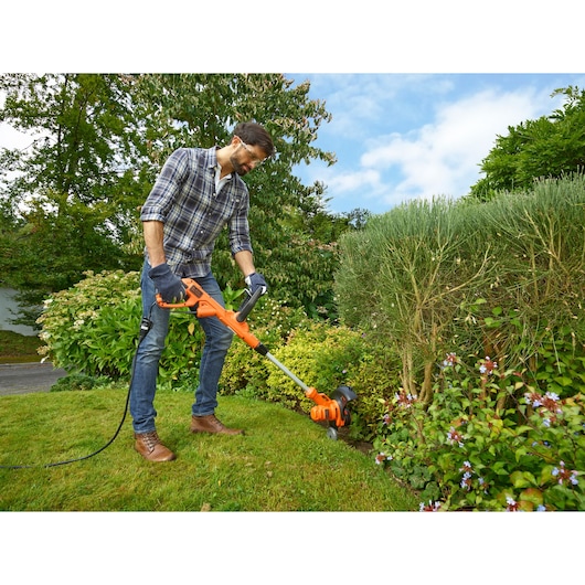 BLACKDECKER BESTA530CM grass trimmer being used in a garden, orange and black design, outdoor setting.