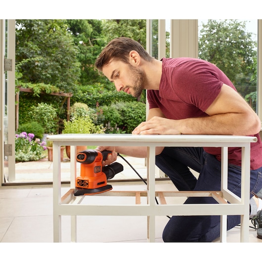 Photo of person using BLACK+DECKER BEW200 detail sander on white table frame indoors near large windows.