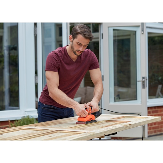 Photo of person using BLACKDECKER BEW220 orange sheet sander on wooden door outdoors.