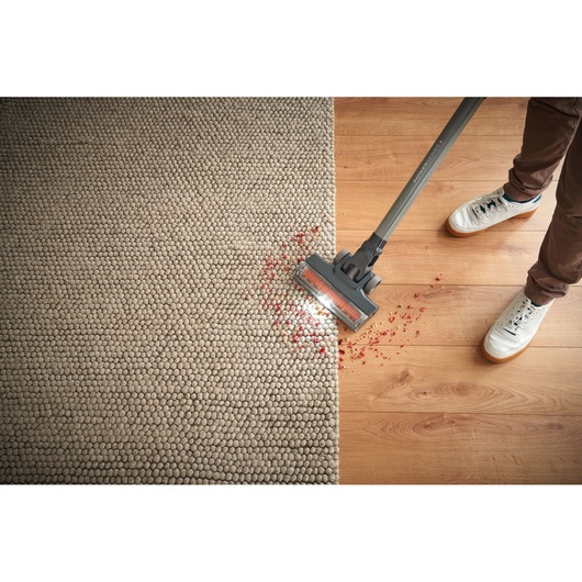 A person is using a BLACK+DECKER cordless vacuum cleaner on a carpet and wooden floor, cleaning up scattered red debris.
