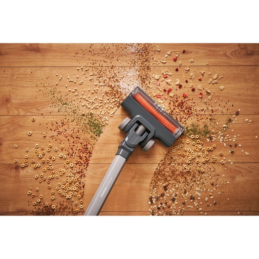 A cordless vacuum cleaner being used on a wooden floor to pick up scattered cereal, herbs, and other small debris.