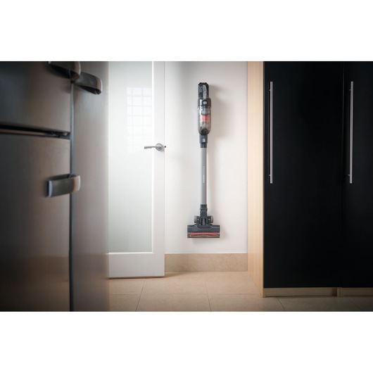 A cordless stick vacuum cleaner mounted on a wall in a modern kitchen, positioned between a refrigerator and a cabinet.