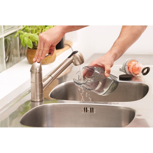 A person rinses a transparent container under a stainless steel kitchen faucet at a sink. Another appliance part is resting near the sink.