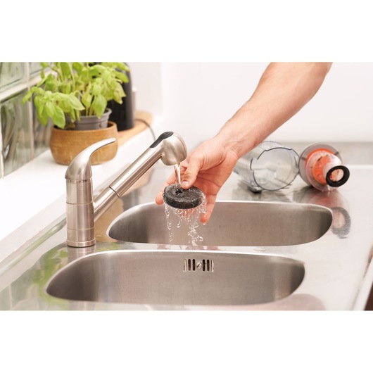 A person's hand is rinsing a vacuum cleaner filter under a kitchen faucet, with vacuum parts on the counter and a potted plant in the background.