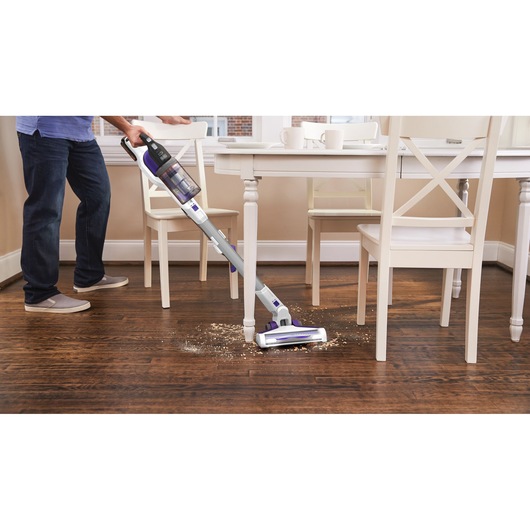BLACKDECKER BHFEV1825CP cordless stick vacuum cleaning crumbs on wood floor near white dining table.