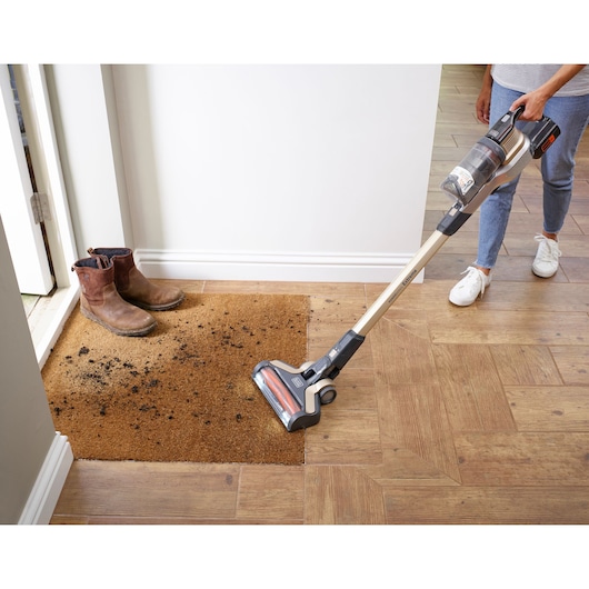 BLACKDECKER BHFEV36B2DW stick vacuum cleaning dirt from a brown doormat near boots, home interior.