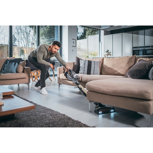BLACKDECKER BHFEV36B2DW cordless vacuum cleaner in use cleaning under beige sofa in modern living room.