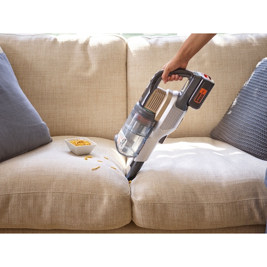 BLACK+DECKER BHFEV36B2DW cordless handheld vacuum cleaning crumbs from beige sofa with grey pillows.