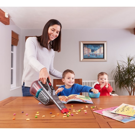 BLACK+DECKER BHHV315B handheld vacuum cleaner used to pick up scattered cereal on wooden dining table in family setting.