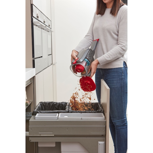 BLACK+DECKER BHHV315B handheld vacuum being emptied into a kitchen trash bin by a person.