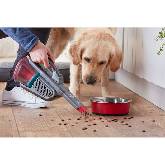 BLACKDECKER BHHV315B handheld vacuum cleaning pet food near a red bowl on wooden floor, side angle.
