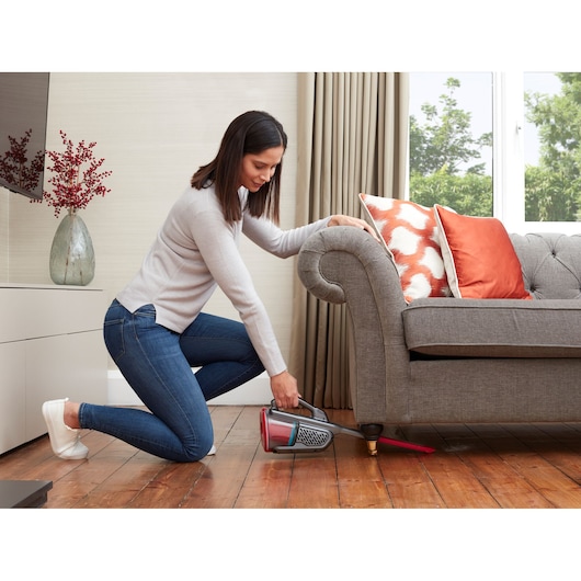 BLACKDECKER BHHV315J handheld vacuum cleaning under a gray sofa with orange cushions in a living room.