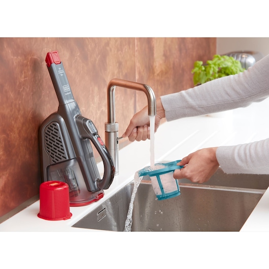 Photo of BLACKDECKER BHHV315J handheld vacuum and filter being cleaned under running water at kitchen sink.