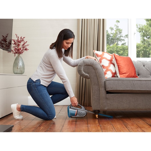 BLACKDECKER BHHV320B handheld vacuum cleaning under sofa in living room with orange and patterned cushions.