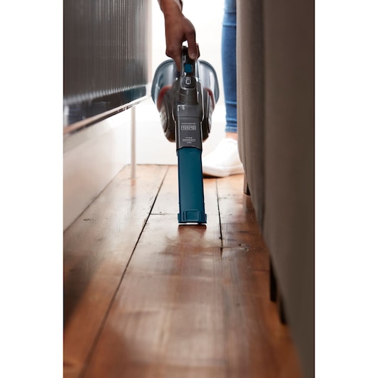 BLACKDECKER BHHV320J handheld vacuum cleaning narrow space beside furniture on wooden floor.